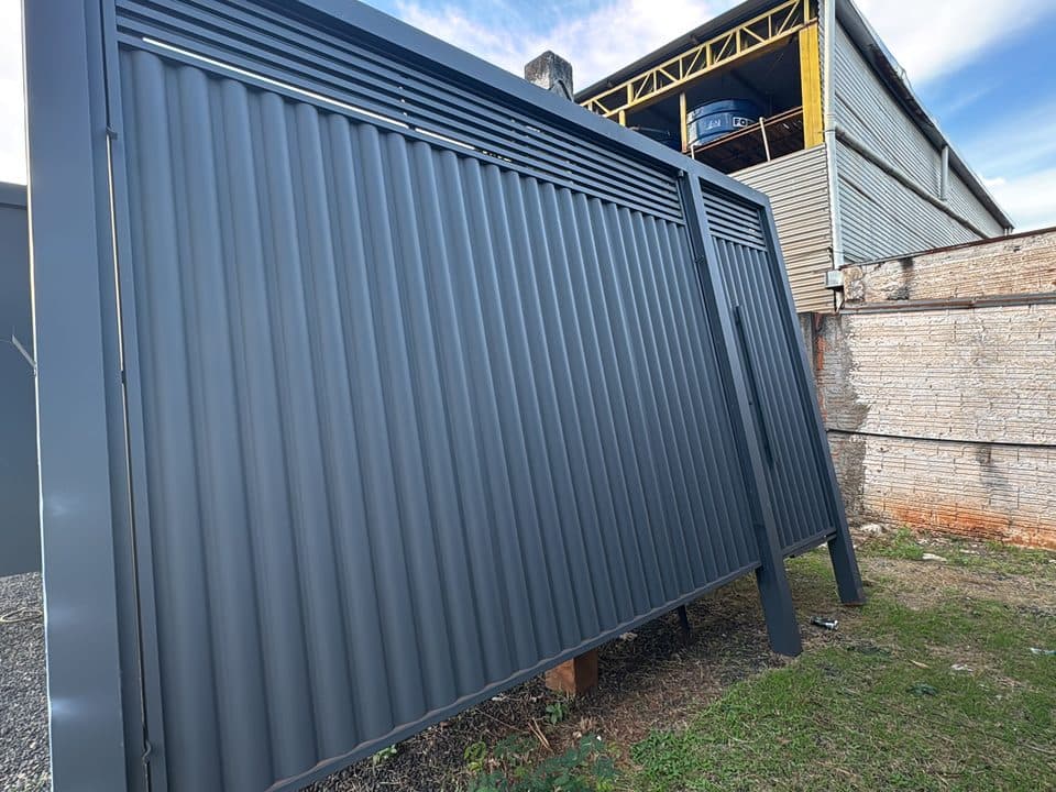 Portão de aço grafite com frisos verticais e ripas no topo, pronto, com o galpão da fábrica ao fundo
